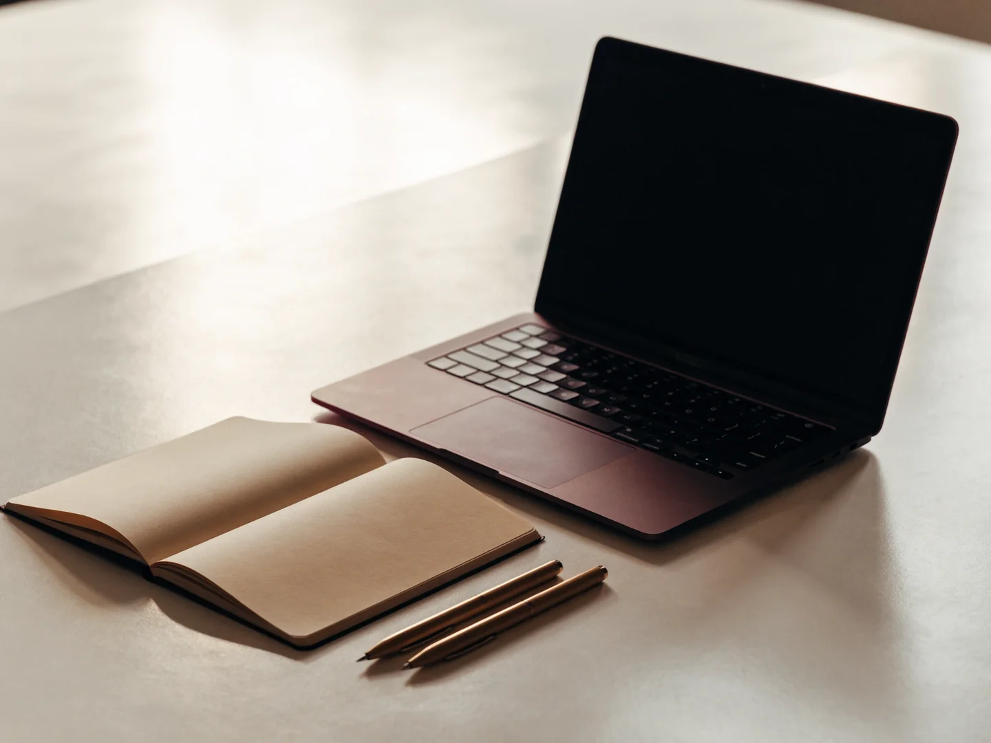 An unbranded laptop, open notebook, and two pens on a light work surface