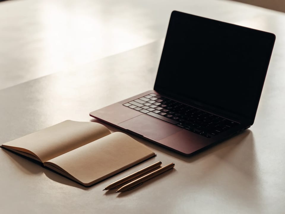 An unbranded laptop, open notebook, and two pens on a light work surface