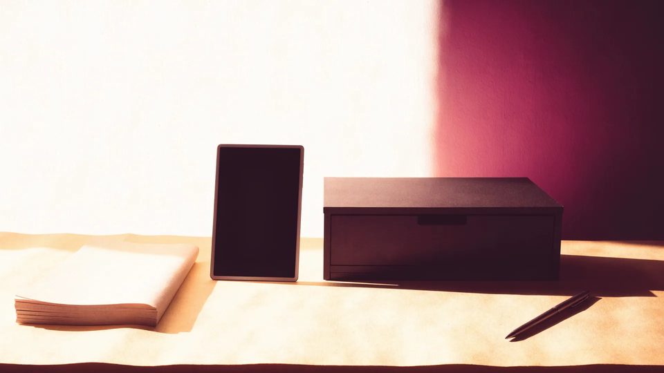 A blank notebook, dark tablet screen, closed storage drawer and pen in warm light.